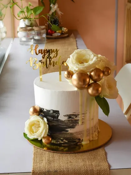 Monochrome with a dash of gold. This elegant birthday cake combines a black-and-white abstract design with a white chocolate drip, fresh white roses, and shiny gold spheres for a chic and modern aesthetic.