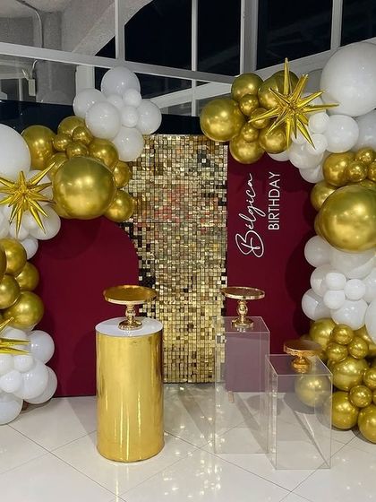 This elegant setup combines a gold shimmer panel with a rich maroon backdrop, framed by a white and gold balloon arch for a very sophisticated look.