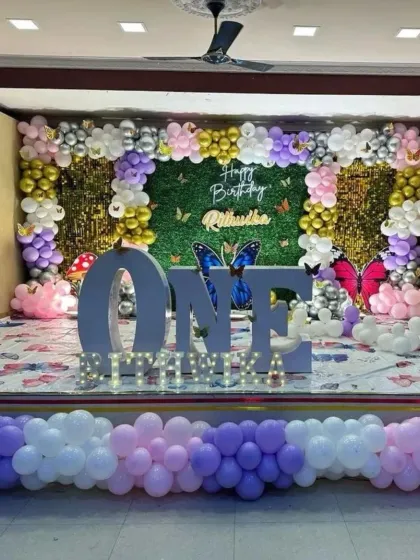 A full-stage butterfly theme decoration for a first birthday. This grand setup includes a large light-up 'ONE' sign, a mixed sequin and green wall backdrop, and butterfly accents throughout the space.