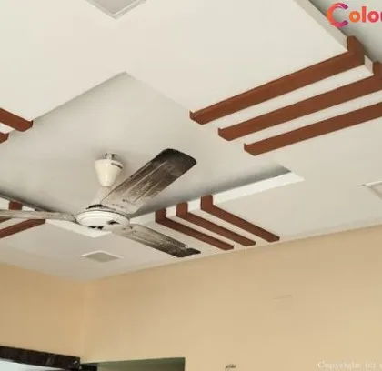 A multi-layered false ceiling featuring clean lines and decorative wooden-finish accents. This design effectively frames the central fan area and adds a sophisticated architectural element to the room.