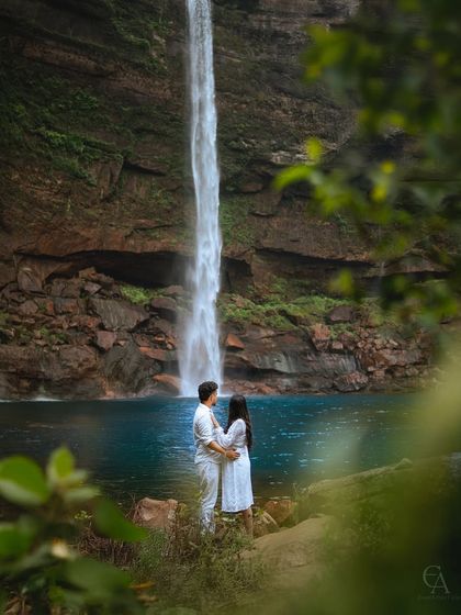 A quiet moment of togetherness, framed by foliage, as they stand before the turquoise waters of the waterfall. This shot feels intimate, as if they are the only two people in the world.