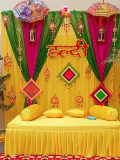 A classic Haldi ceremony backdrop designed for a home setting. This setup features a bright yellow diwan for seating, framed by vibrant yellow, pink, and green drapes, and decorated with traditional phulkari umbrellas and hanging tassels for a festive look.