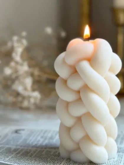 The knotted weave pillar candle, shown lit to create a beautiful, soft glow. The intricate texture casts unique shadows, adding warmth and interest to your room.