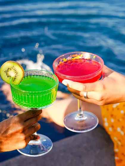 A toast between two people holding colorful cocktails, a green kiwi drink and a red berry one, over sparkling blue water. This image tells a story of friendship and summer fun.