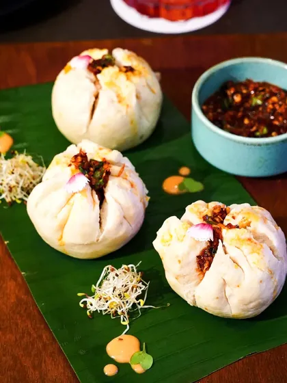 Fluffy, pan-fried bao buns served on a banana leaf with a side of spicy dipping sauce. The close-up shot highlights the soft texture of the buns and the rich filling.