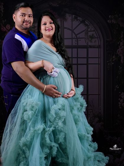 A sweet couple's portrait against a dark floral backdrop. They hold a pair of baby shoes on the baby bump, a simple and touching gesture.