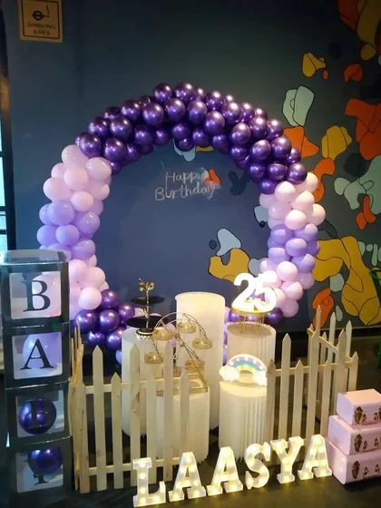 A stylish and compact birthday setup with a purple and lilac circular balloon arch. The decor is personalized with light-up marquee letters for the name "LAASYA" and includes a white picket fence and "BABY" blocks.