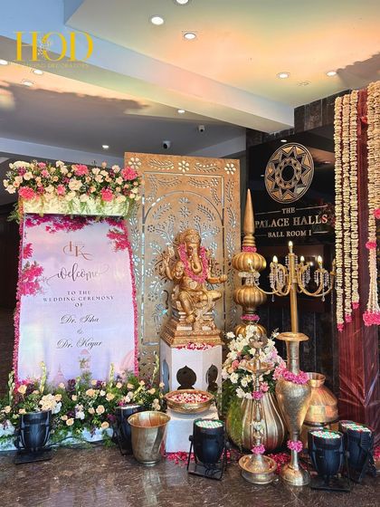 An auspicious welcome corner featuring a divine Ganesh idol, a personalized welcome sign for the couple, and abundant pink and white floral arrangements.