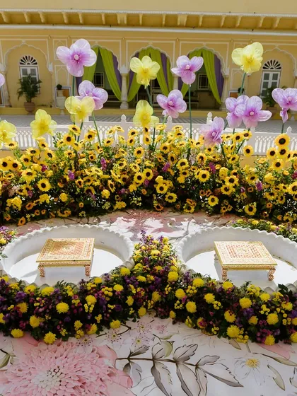The complete Haldi setup for the bride and groom, with two decorated chowkis set amidst a field of vibrant yellow and purple flowers.