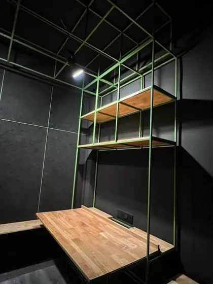 A single workstation within the Hauz Khas micro-studio, showing how the minimal green steel frame and simple wood plank create a functional desk and shelving in a tight space.
