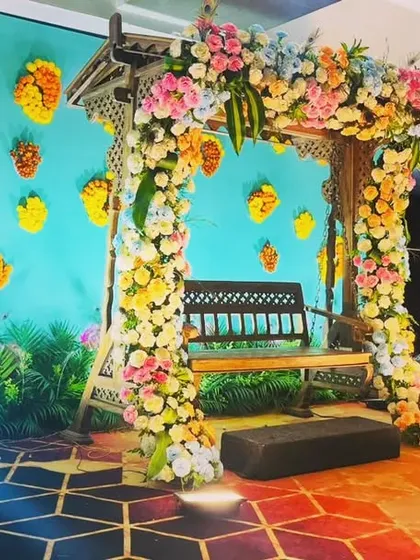 A whimsical and colorful swing setup for a Mehendi or Sangeet. The pastel floral arch and butterfly motifs on the turquoise backdrop create a fun, fairytale-like atmosphere.