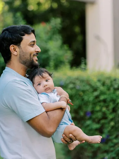 A proud father holding his baby boy outdoors. The natural light and green background create a fresh and lovely family portrait.