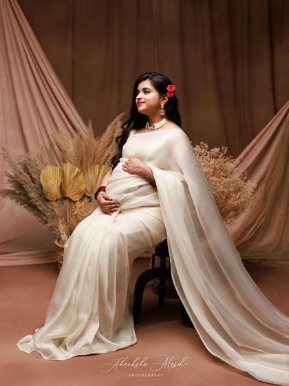 A full-length portrait showing the elegance of a simple white saree. The rustic, earthy backdrop with pampas grass provides a beautiful contrast to the flowing fabric.