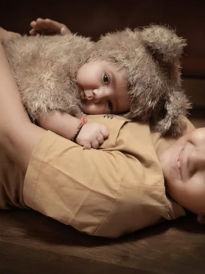 The pure love between brothers. An older brother lies on a wooden floor, gently hugging his baby sibling who is dressed in an adorable fuzzy bear costume. This is a perfect example of our sibling portraits with a newborn.
