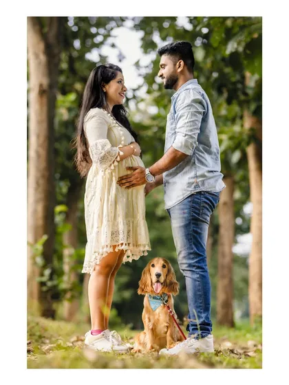 An expecting couple shares a loving gaze in a sunlit park, with their adorable dog sitting patiently at their feet. A perfect portrait of a growing family.