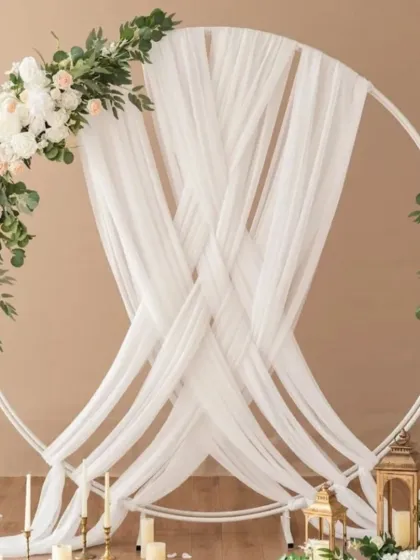 An elegant and minimalist ring ceremony backdrop. This design uses a clean white circular frame, gracefully draped with sheer white fabric and accented with asymmetrical arrangements of white roses and lush green eucalyptus for a sophisticated and modern feel.