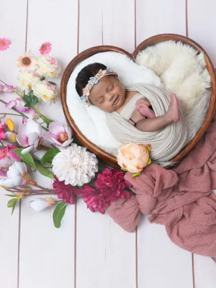 A newborn sleeps soundly in a heart-shaped wooden bowl, nestled on soft white fur and surrounded by an arrangement of pink and white flowers. This composition creates a beautiful and loving portrait.