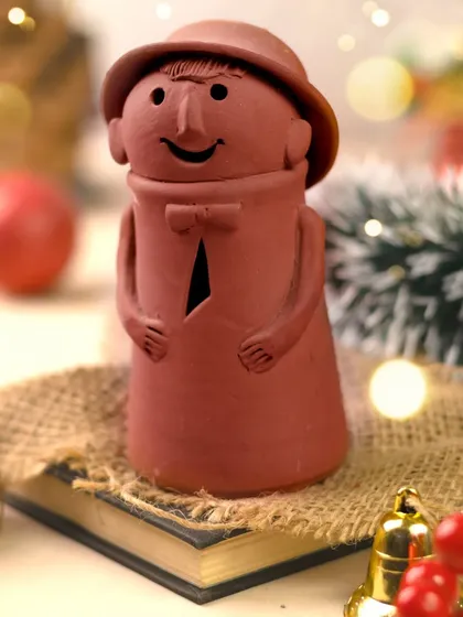 Meet the little clay guardian of holiday cheer. This charming figurine is styled on a book with rustic jute fabric and festive bells, creating a cozy and whimsical Christmas still life.