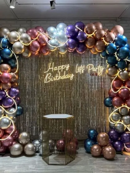 A glamorous party setup with a gold fringe backdrop and a balloon arch made of rich, metallic-colored balloons. The 'Happy Birthday Let's Party' neon sign sets a fun and festive mood.