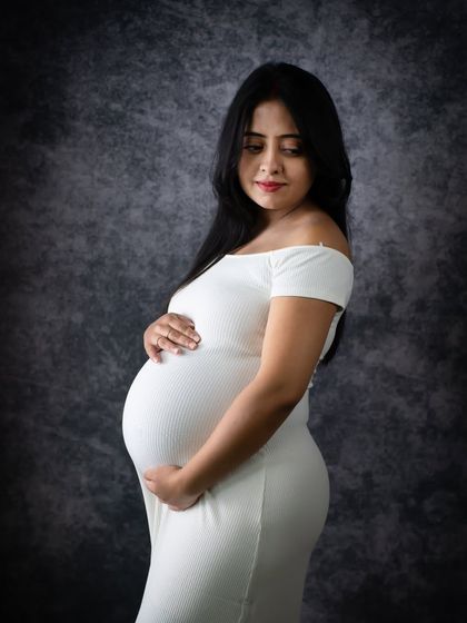 A simple and elegant solo portrait in black and white. The focus is on the beautiful silhouette of the mom-to-be in a classic white dress against a dark backdrop.