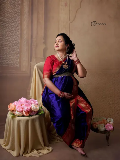 A regal studio portrait of a mother-to-be in a traditional Maharashtrian-style nauvari saree, seated with floral props.