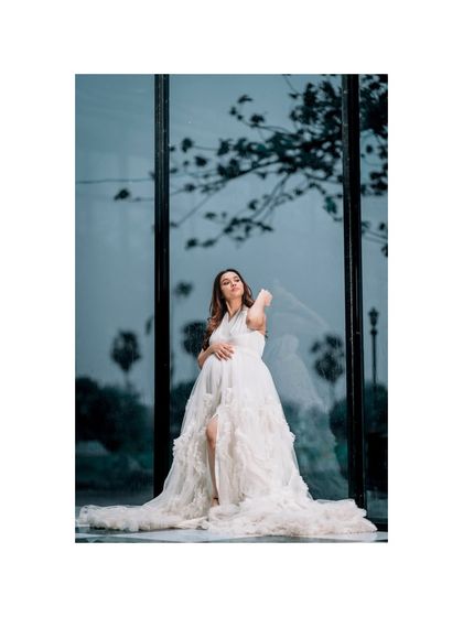 An artistic outdoor shot of a woman in a flowing white maternity gown, looking up towards the sky. The reflection in the glass and the tree branches create a poetic and hopeful image.