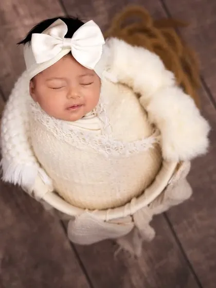 An overhead view of a newborn swaddled in cream-colored fabric inside a small bucket. The large white bow headband adds a touch of elegance to this simple and classic shot.