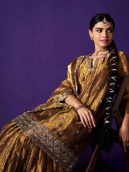 A seated portrait of the model in the gold sharara set, offering another perspective on this stunning celebratory outfit.