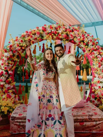A happy couple posing under a vibrant floral arch at their wedding carnival. The arch, filled with bright red, yellow, and pink flowers, served as the perfect photo backdrop.