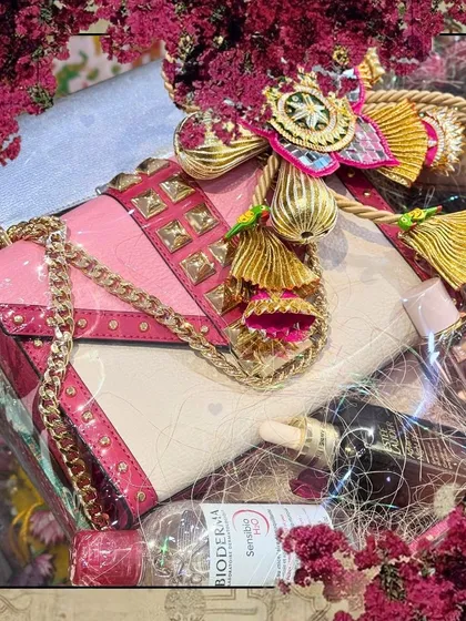 A close-up of a luxury trousseau hamper, framed with a floral border. The vibrant pink handbag and skincare essentials are packaged to create a visually stunning gift.