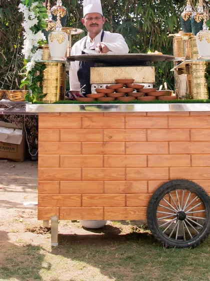 A chef stands ready at a beautifully decorated wooden thela cart. The cart is equipped with elegant serving dishes, ready to provide guests with a unique and interactive food experience.