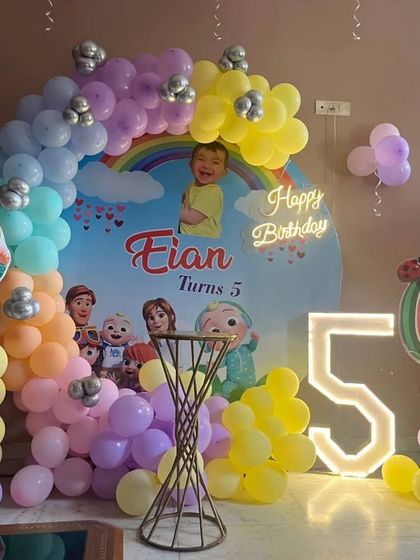 A vibrant Cocomelon party for a 5th birthday. This setup includes a custom printed backdrop, a rainbow-colored balloon arch, character cutouts, and a large, illuminated number '5'.