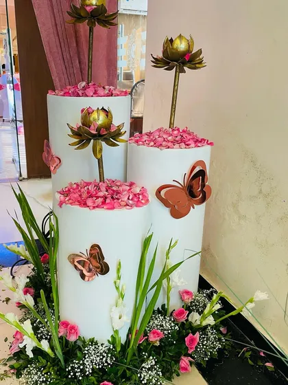 Decorative pillars for a naming ceremony, featuring metallic butterfly cutouts and topped with brass lotus props and fresh rose petals.