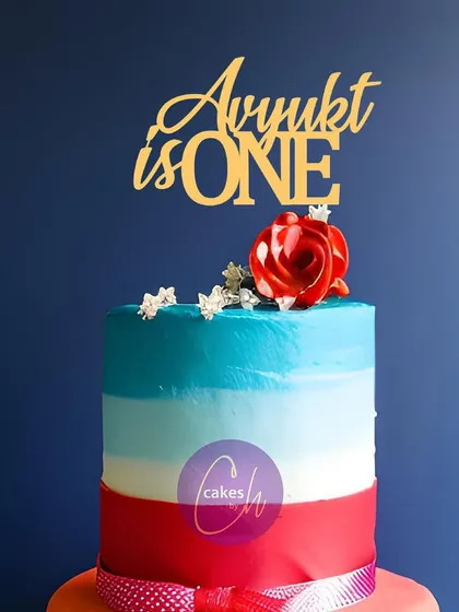 A mockup of the "Avyukt is ONE" topper on a colorful tiered cake, demonstrating how a simple gold topper can complement a vibrant cake design.