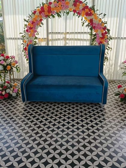 This vibrant floral arch creates a beautiful frame for the couple's seating area. The mix of orange, pink, and white flowers contrasts wonderfully with the royal blue sofa, making it a standout piece for any celebration.