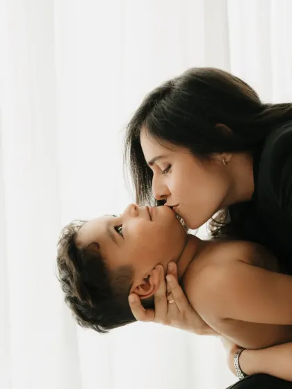 A mother gives her son a gentle kiss. The soft backlighting from the window creates a dreamy and tender mood.
