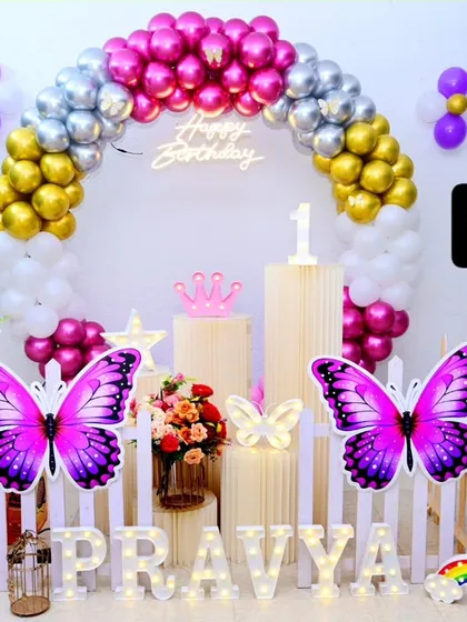 A butterfly garden theme is perfect for a magical first birthday. This setup includes a circular balloon arch in metallic colors, a 'Happy Birthday' neon sign, and large, vibrant butterfly cutouts.