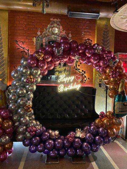 A vibrant and fun birthday setup in a lounge or pub. The balloon ring uses a rich palette of chrome purple, silver, and gold, with butterfly accents and a champagne foil balloon, all centered around a neon sign.