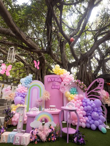 Ayra's rainbow and butterfly first birthday party. This stunning setup under a large tree features pastel rainbow arches, large butterfly wings, and a cascade of colorful balloons.