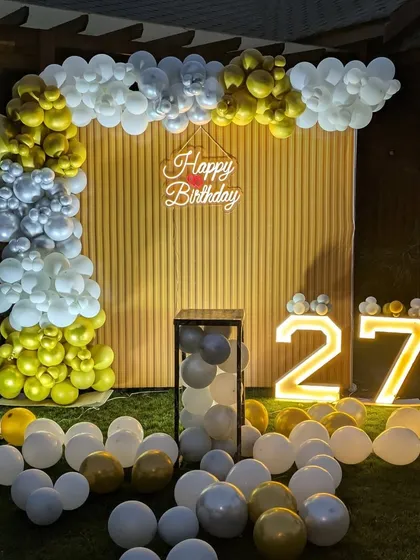 I can turn any outdoor space, like a garden or lawn, into a beautiful party venue. This 27th birthday setup includes a balloon arch against a wooden backdrop, illuminated marquee numbers, and balloons scattered on the grass for a chic, celebratory look.
