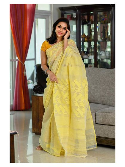 A lifestyle shot featuring a handwoven Dhakai Jamdani saree. The relaxed, seated pose in a home setting makes the product feel warm and approachable.