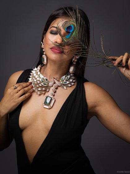 An artistic glamour portrait featuring a grand peacock-inspired necklace. The peacock feather adds a creative and thematic element to the shot.