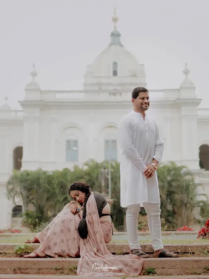 An artistic shot with the groom standing and the bride sitting, set against the backdrop of a grand white palace in Mysore.