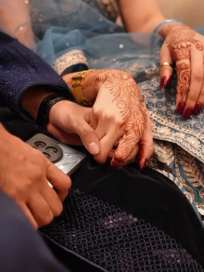 A close-up of the couple's hands, a simple gesture of connection during their engagement ceremony. The intricate mehendi adds a beautiful traditional element.