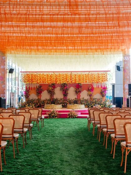 A wide view of a haldi ceremony setup. The stage is decorated with hanging floral arrangements, and the guest seating is arranged on a lush green lawn, creating a fresh and vibrant look.