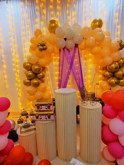 This birthday setup features a peachy-gold and white balloon arch with purple draping. Multiple plinth stands are decorated with props like a mini Ferris wheel and carriage.