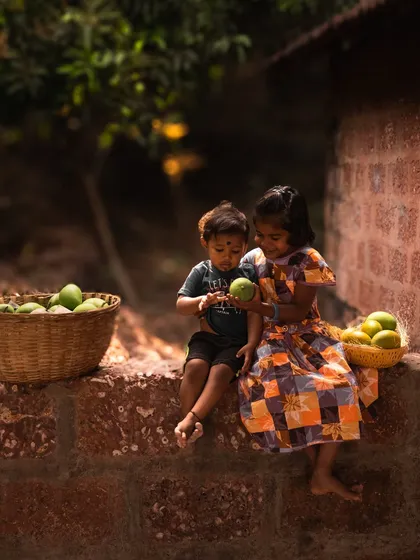 Two children sit on a rustic wall, sharing and inspecting freshly picked mangoes. This image is full of innocence and nostalgia, connecting the product to cherished childhood memories.
