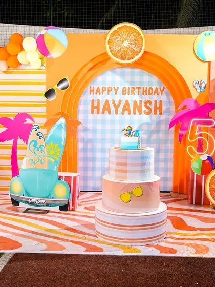 A vibrant, orange-themed summer pool party backdrop. The setup includes surfboards, ice cream props, and a multi-tiered cake, creating a cheerful and festive atmosphere.