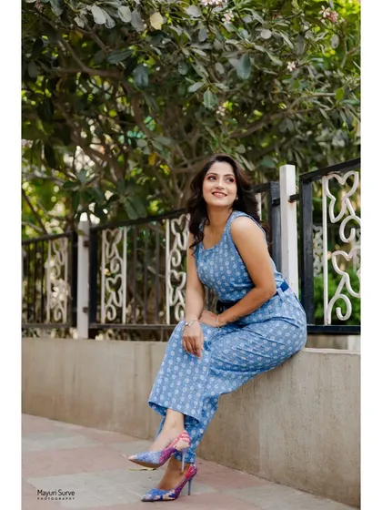 A full-body lifestyle shot for an actress's portfolio. The model is seated casually on a ledge in a stylish blue jumpsuit, creating a confident and modern image.