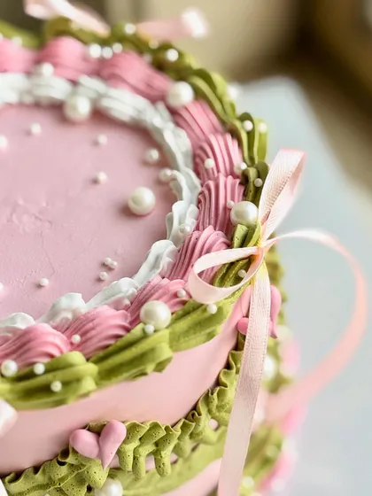 A close-up on the detailed piping work, showing the layers of pink, white, and green buttercream, the scattered pearls, and the soft texture of the satin ribbon.